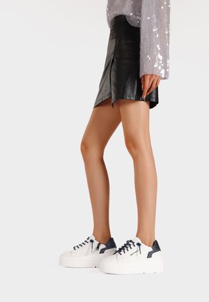 Sneakers bianchi con dettagli blu navy, superficie in pelle texture, punta rotonda e chiusura con lacci, abbinati a una mini gonna nera in pelle.