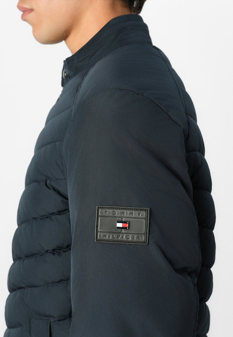 Granatowa kurtka puchowa z pikowanej tkaniny, z zapięciem na zatrzask i gumową naszywką z logo na rękawie. Lekka i dopasowana konstrukcja.