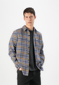 Plaid flannel shirt in blue and brown tones, featuring button-down front, chest pocket, and relaxed fit. Worn over a black T-shirt.