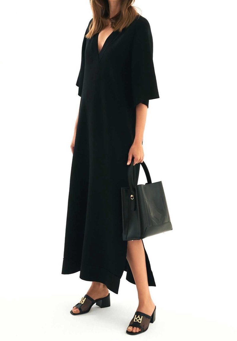 Femme portant une robe midi noire à col en V avec des manches au coude, tenant un sac à main noir et portant des sandales à talons en mesh noir avec des accents dorés.