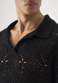 Camisa polo negra de ganchillo con un diseño texturizado y calado, con collar y escote en V, que exhibe patrones intrincados.