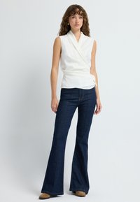 Hvid, ærmeløs indpakningsbluse parret med mørkeblå flared denim jeans. Blusen har et draperet design. Jeansene har detaljer med søm foran og en klassisk pasform.