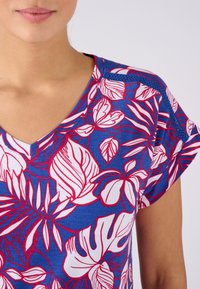 Top floreale con scollo a V che presenta uno sfondo blu con motivi di foglie bianche e rosa, tessuto morbido e maniche corte con accentuazioni in rete.