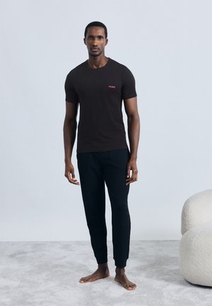 Črna bombažna majica s rdečim logotipom »HUGO«, kombinirana s črnimi joggers hlačami iz teksturirane tkanine. Model stoji na sivem preprogi.