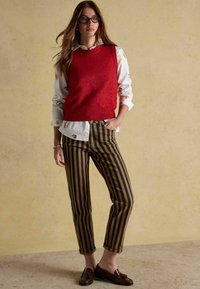 Pulover roșu fără mâneci peste o cămașă albă cu broderie florală, asortat cu pantaloni dungi maro și bleumarin și mocasini maro.