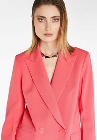 Blazer doppiopetto corallo su misura con rever a spiovente, tasche laterali e bottoni arrotondati. Tessuto dalla texture liscia, design aderente.