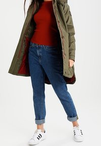 Parka vert olive avec une doublure rouge, haut rouille à côtes, et jeans bleus à manches retroussées. Porté avec des baskets blanches avec des rayures noires.