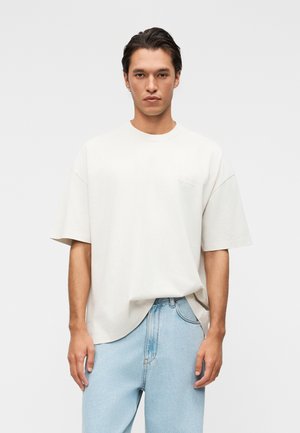 Muž s tmavými vlasmi v nadrozmernom jednofarebnom bielom tričku a svetlomodrých džínsach, stojaci oproti svetlosivému pozadiu.