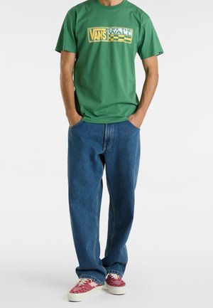 Homme portant un t-shirt Vans vert avec un logo jaune, un jean bleu et des baskets rouges à motif, les mains dans les poches, debout sur un fond blanc.
