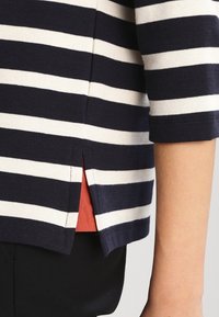 Maglietta a righe blu navy e bianca con maniche a tre quarti, dotata di una fessura laterale e di un sottolivello in corallo a contrasto. Tessuto lavorato a maglia.