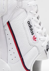 Close-up of white Adidas sneaker with textured leather, red and navy side stripe, and black Adidas logo by white laces on clean background.