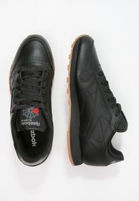 Sorte lærersneakers har en gummisåle, rund tå og teksturerte detaljer. Tungen viser et logo med et stjernedesign.