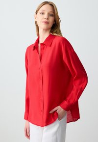 Red linen button-up shirt with a relaxed fit, long sleeves, and a classic collar. Notable for its light, textured fabric and side slits.