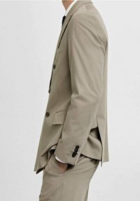 Uomo in abito formale beige con camicia bianca e cravatta nera, in posizione di profilo con una mano in tasca contro uno sfondo semplice.