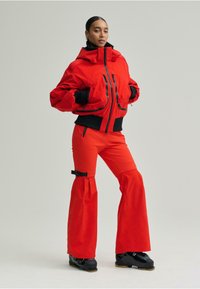 Piros szigetelt dzseki magas gallérral és több elülső zsebbel, bővülő piros nadrággal, oldalsó cipzárokkal és pánt részlettel.