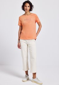 Krátký rukáv, oranžové bavlněné tričko s odraženým textem "Reflection", spojené s bílými zkrácenými kalhotami a šedými teniskami.