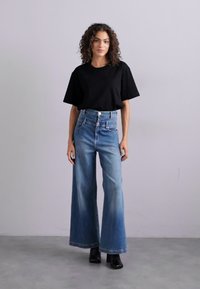 Fekete, rövid ujjú póló magas derekú, bő szárú kék farmernadrággal párosítva. A modell egy semleges háttér előtt áll, fekete bokacsizmát visel.