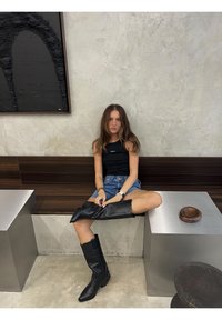 Bottes noires montantes à bout pointu, portées avec des shorts en denim et un débardeur noir sans manches, assises sur un banc en métal contre un mur texturé.