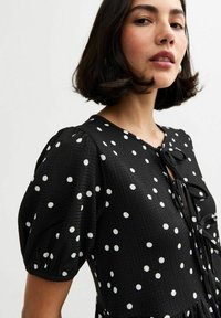 Vestito nero con pois bianchi, tessuto strutturato, maniche a palloncino e dettaglio con nodo sul collo.