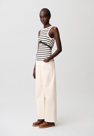 Top senza maniche a righe bianche e nere con dettaglio in pizzo; abbinato a pantaloni larghi color crema e scarpe stringate marroni con lacci decorati.