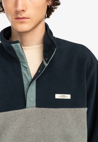 Marinblå och grå fleecepullover med hög krage, utrustad med en ljusblå knappslå och en liten etikett för Element Skateboard Co.