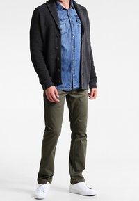 Mörkgrå, ribbade koftan över en ljus denimskjorta, ihop med olivgröna byxor och vita sneakers. Enkla knappar på koftan.