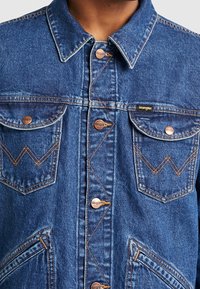 Džínová bunda má tmavě modrou barvu, měděné knoflíky, dvě přední kapsy na hrudi a výrazné oranžové prošití ve vzoru cikcak.