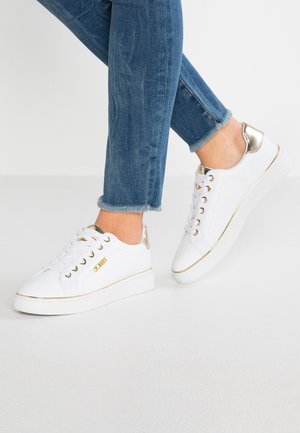 Pies con zapatillas blancas de caña baja con detalles y cordones dorados, combinadas con vaqueros azules desgastados hasta el tobillo sobre un suelo claro.