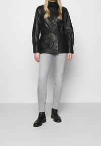 Persona vestida con una chaqueta de cuero negra con cinturón, jeans skinny gris claro, botas negras hasta el tobillo, de pie contra un fondo blanco liso.