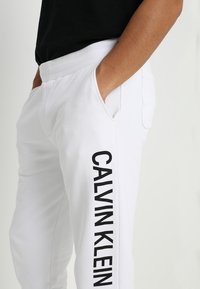 Białe spodnie dresowe wykonane z miękkiej tkaniny z czarnym nadrukiem "CALVIN KLEIN" wzdłuż boku, wyposażone w elastyczny pas i boczne kieszenie.