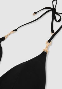 Top bikini nero con connettori metallici rettangolari dorati e sottili spalline regolabili annodate a fiocco su uno sfondo grigio chiaro.