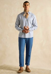 Svītraina pogas krekls zilos un baltos toņos, savienots ar zilām biksēm. Akcentēts ar gaiši brūnu apkakles šalli un brūniem loaferiem.