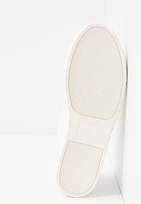 Semelle de chaussure blanche avec des rainures horizontales et un logo circulaire. Le matériau semble caoutchouteux avec une finition lisse et mate et des bords arrondis.