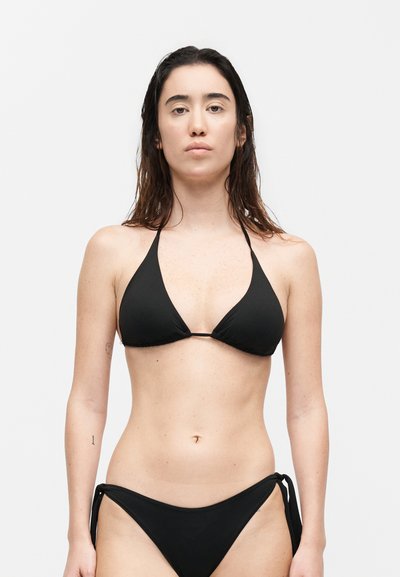 Νεαρή γυναίκα με μακριά σκούρα νωπά μαλλιά, όρθια, φορώντας ένα απλό μαύρο δεμένο μπικίνι πάνω σε λευκό φόντο.