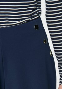 Navy blue trousers with a smooth texture, featuring three metallic black buttons on the side and paired with a striped long-sleeve top.
