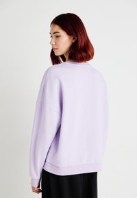 Felpa lilla con spalle scese e polsini a costine, caratterizzata da una vestibilità rilassata e da una texture morbida. Indossata con pantaloni neri.
