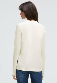 Cremefarbenes Langarmshirt aus Baumwolle mit rundem Halsausschnitt und seitlichem Etikett, kombiniert mit blauen Denim-Jeans.