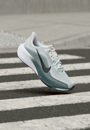 Scarpa da corsa Nike grigio chiaro e verde con swoosh nero, in aria sopra una strisce pedonali su una strada asfaltata.