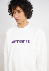 Sweatshirt blanc à col rond et manches longues, avec un logo "carhartt" brodé violet bien en évidence sur la poitrine.