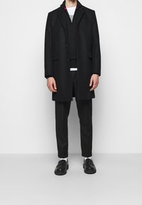Abrigo de lana negro con cierre de botón, bolsillo de parche, superpuesto sobre un suéter oscuro y pantalones, combinado con zapatos negros y calcetines blancos.