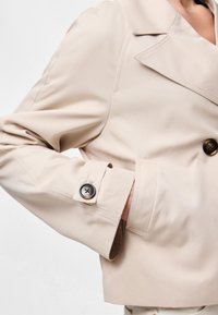 Cappotto beige con bottoni tartarugati, mano in tasca e ampio rever, su parte superiore del corpo e braccio di una persona.