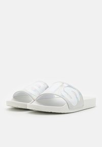 Sandales à glissière blanches avec une bride irisée brillante ornée de lettres surdimensionnées. La semelle est plate et lisse, offrant un confort décontracté.