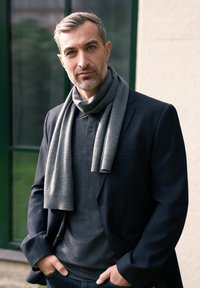 Mand i en mørk blazer over en mørk polo-shirt, med et gråt strikket tørklæde hængende om halsen, der står mod en lys væg.