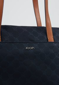 Sac cabas noir avec texture subtilement texturée, poignées en cuir marron et logo métallique "JOOP!" sur le devant.