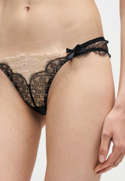 Femme portant une culotte en dentelle noire avec des motifs floraux et un nœud sur le côté, montrant le bas-ventre et le haut des cuisses.