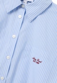 Camicia azzurra chiaro con righe verticali bianche e blu, dotata di taschino sul petto con testo ricamato rosso e colletto classico.