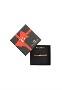 Bracciale dorato con un emblema circolare, presentato in una scatola nera con un nastro rosso e un fiocco decorato con un dettaglio a forma di ferro di cavallo.