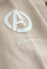 Heled beezid kangas, millel on tekstuuriga, tõstetud valge logo "A" ja allpool selge ja julge kirjaga tekst "THE AVENGERS".