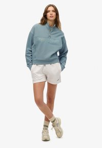  Lichtblauw sweatshirt met kwart rits en het logo "SUPERDRY", gecombineerd met witte shorts. Model draagt beige sneakers en gestreepte groene sokken.