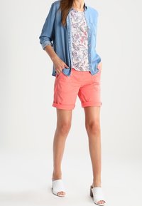 Chemise bleu clair à boutons, haut blanc à motif floral, shorts corail, mules ouvertes blanches. Tenue décontractée avec shorts retroussés et poches avant.